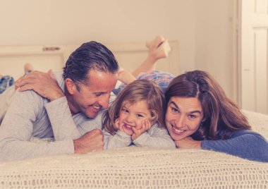 Güzel mutlu aile portresi. Rahatlıyor ve evde yatakta birlikte vakit geçiriyorlar. Anne, baba ve küçük tatlı kız sabah öpüşüp kaynaşırken eğleniyorlar..