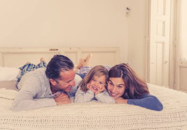 Güzel mutlu aile portresi. Rahatlıyor ve evde yatakta birlikte vakit geçiriyorlar. Anne, baba ve küçük tatlı kız sabah öpüşüp kaynaşırken eğleniyorlar..