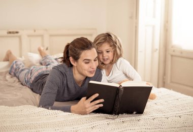 Genç anne ve küçük tatlı kız birlikte kitap okuyorlar. Mutlu anne ve tatlı sarışın kızın yatakta masal okurken eğlendikleri hayat tarzı. Mutlu aile, ebeveynlik ve eğitim konseptinde.