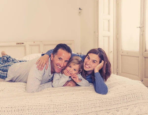 Lifestyle portrait of beautiful happy family relaxing and spending time together in bed at home. Mother, father, and cute toddler daughter having fun kissing hugging and bonding in the morning.