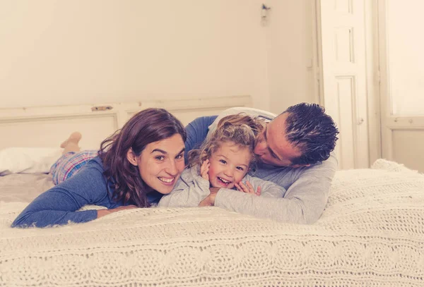 Lifestyle portrait of beautiful happy family relaxing and spending time together in bed at home. Mother, father, and cute toddler daughter having fun kissing hugging and bonding in the morning.