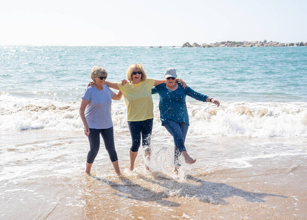 Прекрасная группа из трех пожилых женщин в возрасте 60 лет, гуляющих и веселящихся на пляже. Зрелые женщины смеются и пинают воду улыбаясь игриво наслаждаясь пенсионным образом жизни и дружбы
.