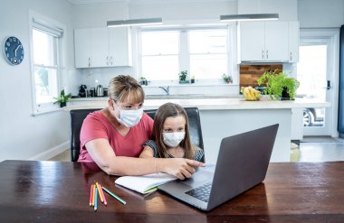 Coronavirus salgını. Kilitlenme ve okul kapanması. Anne, yüz maskeli sıkılmış kızının evde online ders çalışmasına yardım ediyor. COVID-19 salgını çocukları ve öğretmenleri çevrimiçi öğrenmeye zorluyor.