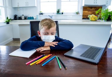 Coronavirus salgını. Kilitlenme ve okul kapanması. Yüz maskeli okul çocuğu evde sıkılmış ve depresif hissederek online eğitim dersleri izliyor. COVID-19 salgını çocukları çevrimiçi öğrenmeye zorluyor.