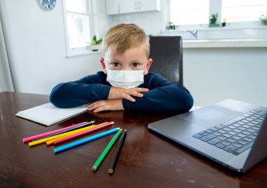 Coronavirus salgını. Kilitlenme ve okul kapanması. Yüz maskeli okul çocuğu evde sıkılmış ve depresif hissederek online eğitim dersleri izliyor. COVID-19 salgını çocukları çevrimiçi öğrenmeye zorluyor.