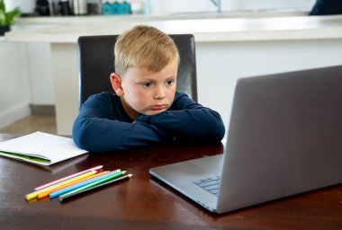 Coronavirus salgını. Kilitlenme ve okul kapanması. Yüz maskeli okul çocuğu evde sıkılmış ve depresif hissederek online eğitim dersleri izliyor. COVID-19 salgını çocukları çevrimiçi öğrenmeye zorluyor.