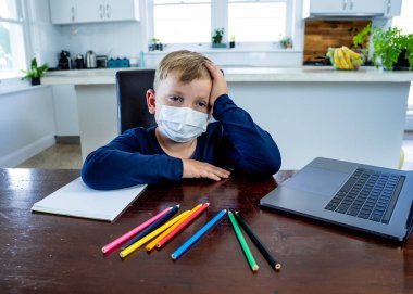Coronavirus salgını. Kilitlenme ve okul kapanması. Yüz maskeli okul çocuğu evde sıkılmış ve depresif hissederek online eğitim dersleri izliyor. COVID-19 salgını çocukları çevrimiçi öğrenmeye zorluyor.