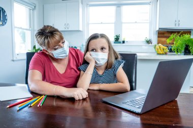 Coronavirus salgını. Kilitlenme ve okul kapanması. Anne, yüz maskeli sıkılmış kızının evde online ders çalışmasına yardım ediyor. COVID-19 salgını çocukları ve öğretmenleri çevrimiçi öğrenmeye zorluyor.