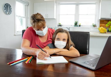 Coronavirus salgını. Kilitlenme ve okul kapanması. Anne, yüz maskeli sıkılmış kızının evde online ders çalışmasına yardım ediyor. COVID-19 salgını çocukları ve öğretmenleri çevrimiçi öğrenmeye zorluyor.