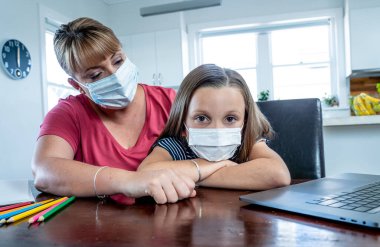 Coronavirus salgını. Kilitlenme ve okul kapanması. Anne, yüz maskeli sıkılmış kızının evde online ders çalışmasına yardım ediyor. COVID-19 salgını çocukları ve öğretmenleri çevrimiçi öğrenmeye zorluyor.