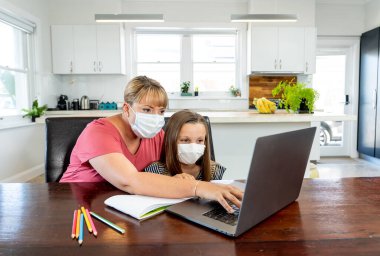 Coronavirus salgını. Kilitlenme ve okul kapanması. Anne, yüz maskeli sıkılmış kızının evde online ders çalışmasına yardım ediyor. COVID-19 salgını çocukları ve öğretmenleri çevrimiçi öğrenmeye zorluyor.