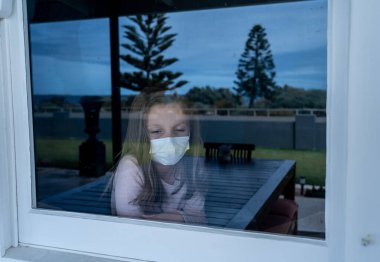 COVID-19 Lockdown. Depressed lonely little girl with face mask looking through the window during quarantine. Sad sick child in self isolation at home. Coronavirusu outbreak and children mental health. clipart