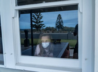 COVID-19 tecrit. Karantina sırasında yüz maskeli depresyondaki küçük kız pencereden dışarı bakıyor. Evde kendini izole etmiş üzgün hasta bir çocuk. Coronavirusu salgını ve çocuk sağlığı.