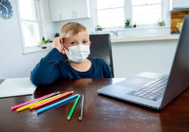Coronavirus salgını. Kilitlenme ve okul kapanması. Yüz maskeli okul çocuğu evde sıkılmış ve depresif hissederek online eğitim dersleri izliyor. COVID-19 salgını çocukları çevrimiçi öğrenmeye zorluyor.