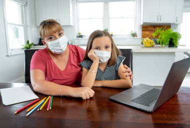 Coronavirus salgını. Kilitlenme ve okul kapanması. Anne, yüz maskeli sıkılmış kızının evde online ders çalışmasına yardım ediyor. COVID-19 salgını çocukları ve öğretmenleri çevrimiçi öğrenmeye zorluyor.