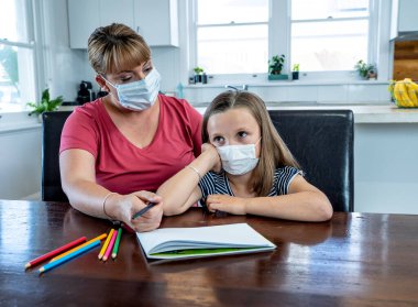 Coronavirus salgını. Kilitlenme ve okul kapanması. Anne, yüz maskeli sıkılmış kızının evde online ders çalışmasına yardım ediyor. COVID-19 salgını çocukları ve öğretmenleri çevrimiçi öğrenmeye zorluyor.