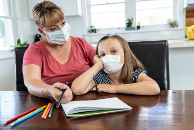 Coronavirus salgını. Kilitlenme ve okul kapanması. Anne, yüz maskeli sıkılmış kızının evde online ders çalışmasına yardım ediyor. COVID-19 salgını çocukları ve öğretmenleri çevrimiçi öğrenmeye zorluyor.