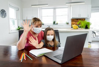 Coronavirus salgını. Kilitlenme ve okul kapanması. Anne, yüz maskeli sıkılmış kızının evde online ders çalışmasına yardım ediyor. COVID-19 salgını çocukları ve öğretmenleri çevrimiçi öğrenmeye zorluyor.