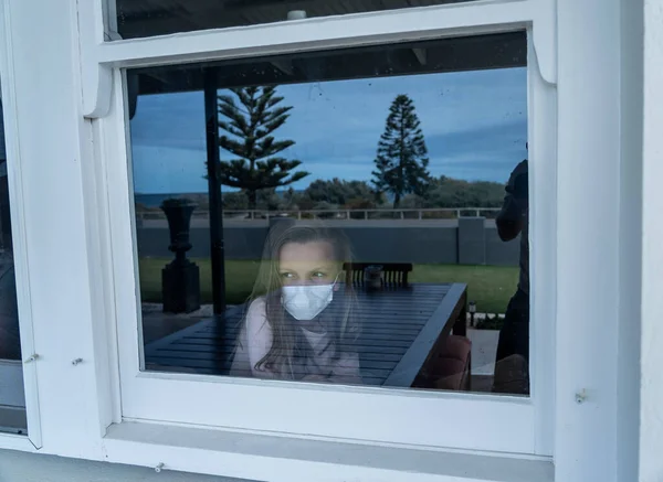 COVID-19 Lockdown. Depressed lonely little girl with face mask looking through the window during quarantine. Sad sick child in self isolation at home. Coronavirusu outbreak and children mental health.