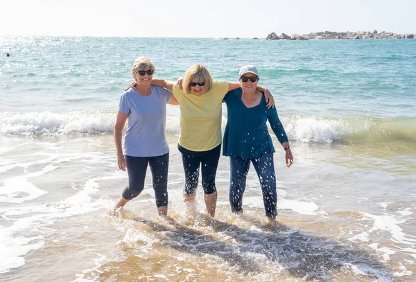 由三位60多岁的老年退休妇女组成的可爱的小组在沙滩上散步和玩乐 成熟女性笑着踢水嬉笑着享受退休生活和友谊 — 图库照片