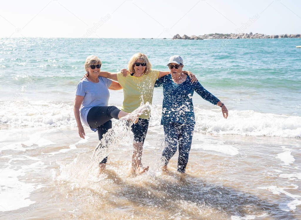 Lovely group of senior girl friends on their 60s walking and having fun splashing water on sea. Three mature healthy retired females Laughing and enjoying retirement and outdoors active lifestyle.