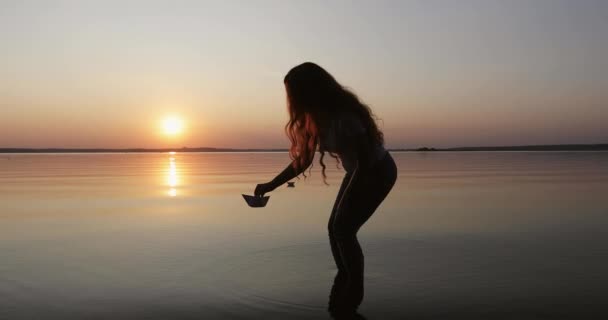 Sylwetka dziewczyny z długimi kręconymi włosami kładącej papierową łódkę na wodzie. — Wideo stockowe