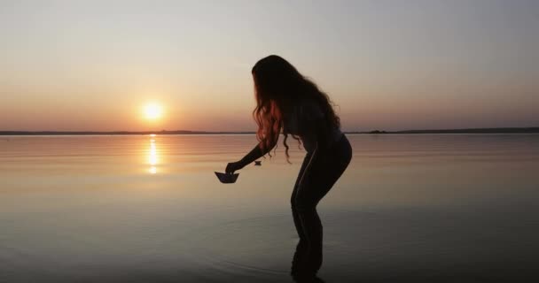 Silhouette of a girl with long curly hair putting down paper boat on the water. — ストック動画