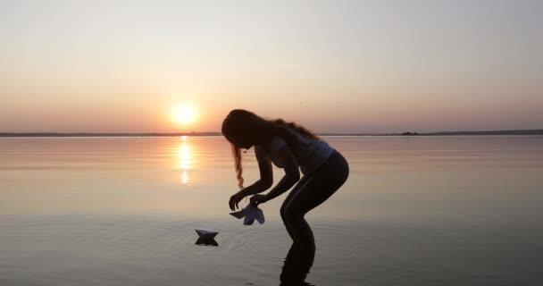 Silhouette d'une fille aux longs cheveux bouclés laissant quelques bateaux en papier aller sur l'eau . — Video