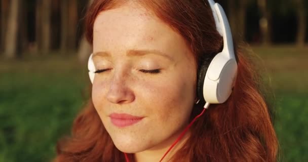 Close up face of redhead freckled girl with white headphones is listening songs and smiling. — Stock Video