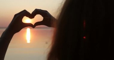 Close up head and hands. Silhouette of a girl making a heart with hands at sunset.