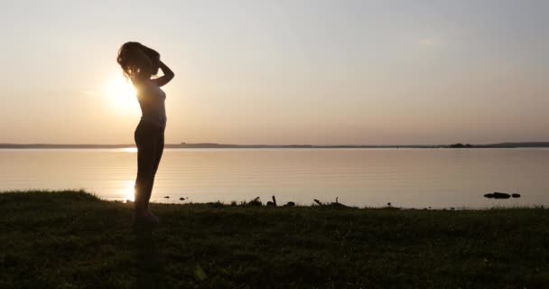 Молодая женщина с длинными вьющимися рыжими волосами стоит на берегу озера на закате . — стоковое видео