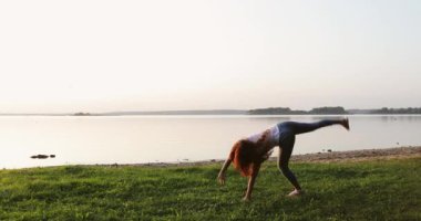 Young sporty woman with long curly red hair doing excersises on shore of the lake.