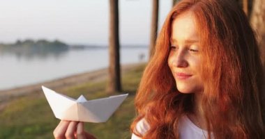Portrait of a girl with red hair holding a white paper boat in hand.