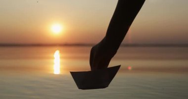 Silhouette of a human hand letting go a paper boat in water of the sea or lake at sunset. T