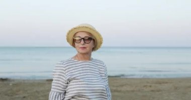 Hasır şapkalı neşeli yaşlı kadın sabahın erken saatlerinde deniz sahilinde yürüyor..