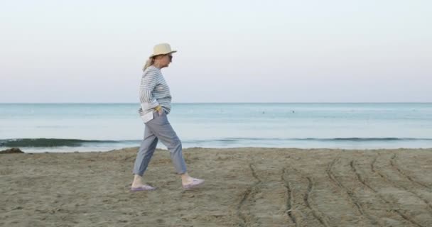 Die fröhliche ältere Frau mit Strohhut, die frühmorgens bei Sonnenaufgang am Meeresstrand spaziert. — Stockvideo