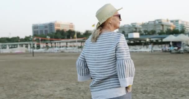 Wesoła starsza kobieta w słomkowym kapeluszu spacerująca po morskiej plaży wczesnym rankiem o wschodzie słońca. — Wideo stockowe
