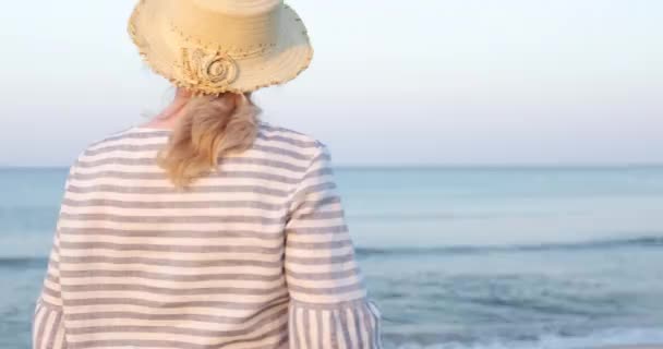 Starsza kobieta w słomkowym kapeluszu, stojąca wczesnym rankiem na plaży morskiej. — Wideo stockowe