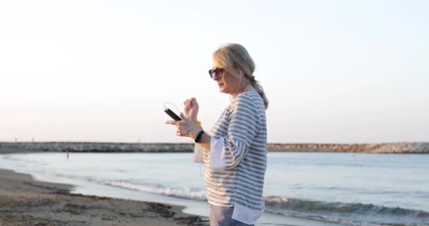 カメラフライ周り60歳の女性スリムイヤフォンで音楽を楽しむ. — ストック動画
