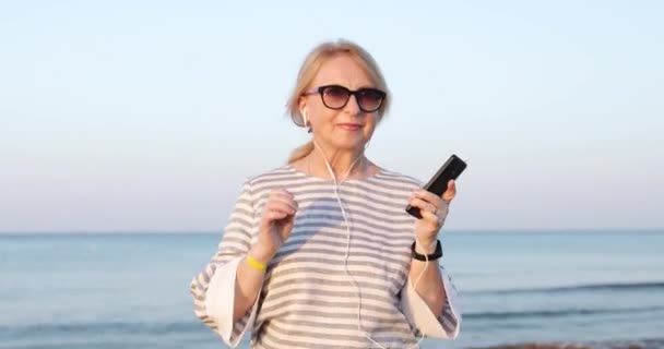 Blond äldre kvinna i solglasögon promenader och danser på en havsstrand lyssna en musik på mobiltelefon. — Stockvideo