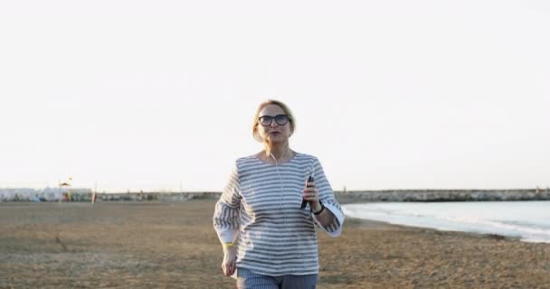 Блондинка літня жінка в сонцезахисних окулярах бігає і танцює на морському піщаному пляжі . — стокове відео