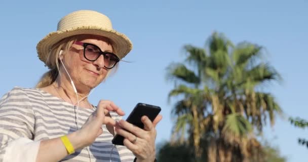Blond bejaarde vrouw in zonnebril en strohoed kiezen van een lied op mobiele telefoon. — Stockvideo