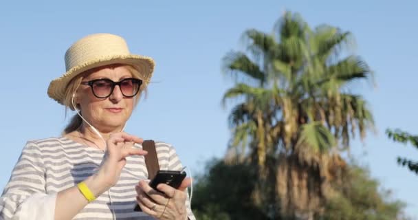 Blondýny starší žena v slunečních brýlích a slamáku poslech hudby na mobilním telefonu ve sluchátkách. — Stock video