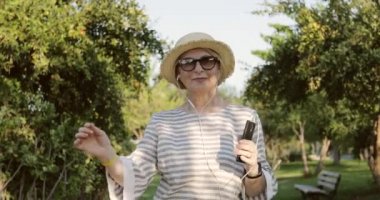 Güneş gözlüklü, şapkalı, kulaklıklı, cep telefonuyla müzik dinleyen sarışın yaşlı bir kadın..