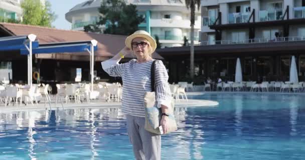 The cheerful elderly woman in straw hat walking by the pool at the hotel. — Stock Video