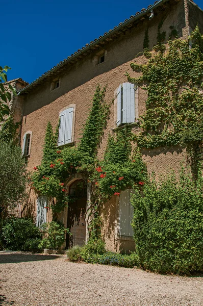 Güneşli mavi gökyüzü ve çiçeklerle Menerbes Köyü, bir avluya bakan tipik taş ev görünümünü. 