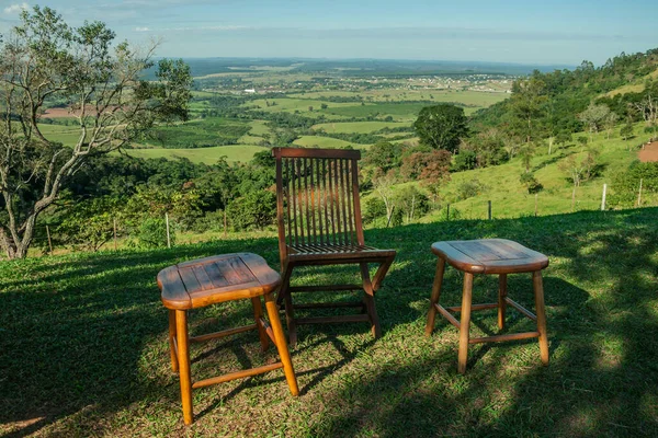 Wooden stool and chair in the shade in front of green valley on hilly landscape covered by forest and meadow near Pardinho. A small rural village in the countryside of Sao Paulo State.