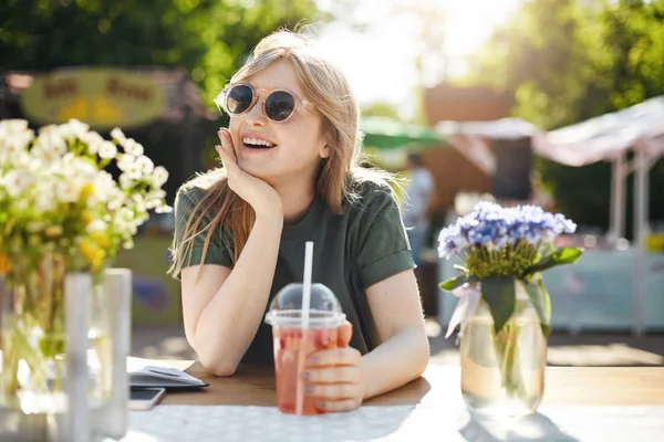 Porträt einer entzückenden jungen Frau, die Limonade im von Blumen umgebenen Park trinkt und eine Brille trägt, die von ihrer zukünftigen Karriere träumt. — Stockfoto