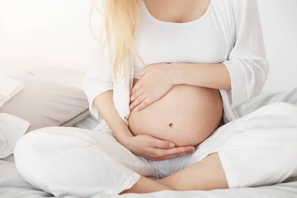 Jovem grávida em breve será mãe tocando sua barriga sentada em seu quarto à tarde. Conceito de gravidez. Chave alta . — Fotografia de Stock
