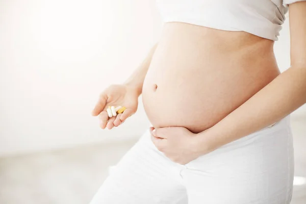Close up of soon to be happy mother holding belly and prenatal pills and vitamins and minerals waiting for a healthy baby. Prenatal concept. High key. — Stock Photo, Image